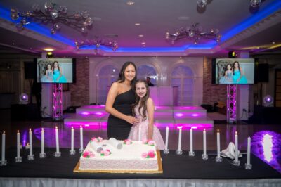 Two people, a woman in a black dress and a girl in a pink dress, stand smiling behind a large rectangular cake with pink and white decorations at an event hall in NY, enjoying entertainment services Suffolk and Nassau County, NY events love.