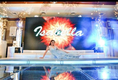 A young woman in a light blue gown sits on a shiny stage in NY, in front of a large screen displaying "Isabella" with a colorful burst design, surrounded by bright lights and spark effects, showcasing top entertainment services Suffolk and Nassau County.