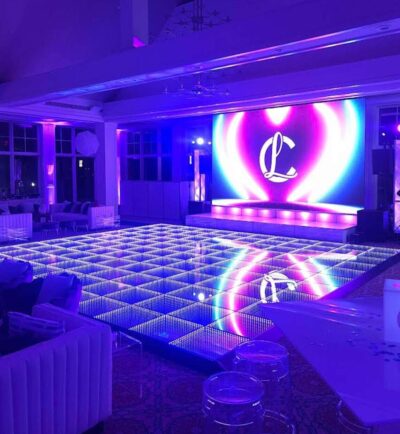 A large, modern event space in NY with a glowing LED dance floor and a vibrant screen displaying a cursive "L" against a colorful, heart-shaped background. The room features purple and blue accent lights and offers top DJ services in Suffolk and Nassau County, NY.