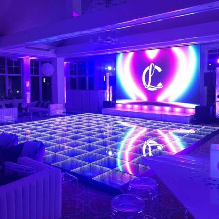 A large, modern event space in NY with a glowing LED dance floor and a vibrant screen displaying a cursive "L" against a colorful, heart-shaped background. The room features purple and blue accent lights and offers top DJ services in Suffolk and Nassau County, NY.