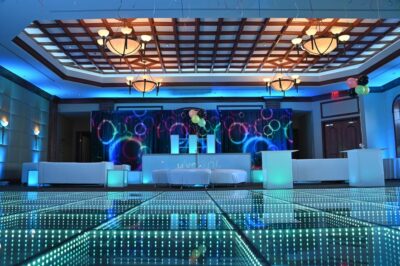 A modern, elegant event venue featuring a glowing LED dance floor, white lounge furniture, colorful balloons, decorative lighting, and a vibrant neon backdrop. Perfect for entertainment services Suffolk and Nassau County, NY. Chandeliers hang from the coffered ceiling.