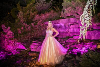 A woman in a sparkling, light-colored ball gown poses outdoors at night, surrounded by rocks and greenery illuminated with pink and purple lights—an enchanting NY,NY scene perfect for events with top dj services Suffolk and Nassau County.