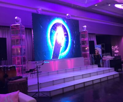 A stage with a large, illuminated screen displays a glowing, circular blue and white graphic. Lit by purple lights with musical instruments and speakers set up, it showcases top entertainment services Suffolk and Nassau County, NY. A person stands to the side.