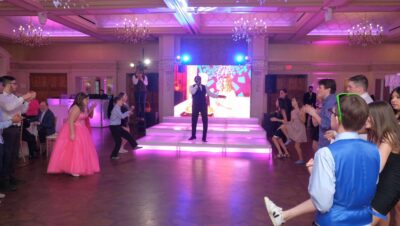 A group dressed formally gathers on a dance floor in a purple-lit hall, enjoying a performer on stage. One girl in pink looks ready to dance, highlighting the vibrant dj services Suffolk and Nassau County offer for unforgettable events.