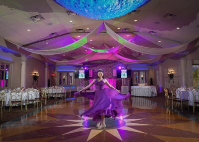 A woman in a flowing purple dress twirls gracefully on a star-patterned floor in a banquet hall illuminated by colorful lighting, with draped fabric overhead—an enchanting scene brought to life by top entertainment services Suffolk and Nassau County.