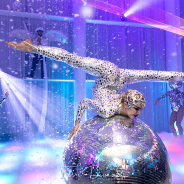 A performer in a sparkling bodysuit does a split on a giant disco ball, surrounded by dancers with mirror ball helmets and colorful lights—NY, NY entertainment services for Suffolk and Nassau County bring this futuristic, vibrant scene to life.