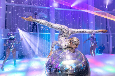 A performer in a sparkling bodysuit does a split on a giant disco ball, surrounded by dancers with mirror ball helmets and colorful lights—NY, NY entertainment services for Suffolk and Nassau County bring this futuristic, vibrant scene to life.