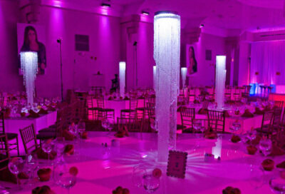 A banquet hall decorated with tall, illuminated crystal centerpieces on round tables, bathed in pink-purple lighting. Perfect for events enhanced by top entertainment services Suffolk and Nassau County, NY. A festive and elegant atmosphere awaits.