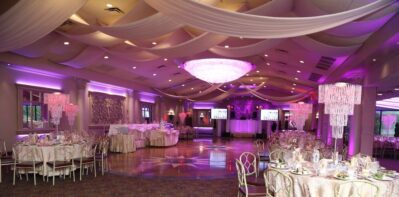 Elegant banquet hall decorated for an event, featuring round tables with gold chairs, draped white fabric on the ceiling, large chandeliers, and purple lighting. A dance floor is centered in the room—perfect for dj services Suffolk and Nassau County, NY.