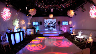 A modern NY,NY event lounge with illuminated furniture, a central dance floor with colorful swirling light patterns, white décor, floral arrangements, and a large digital screen—perfect for parties needing DJ services in Suffolk and Nassau County.