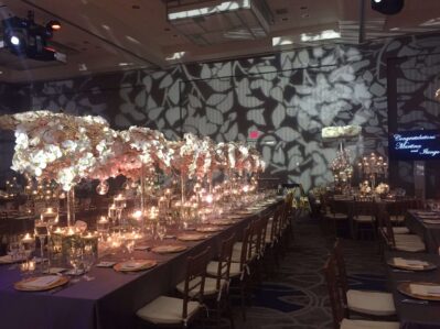 Elegant banquet hall in NY, NY, with long tables adorned by tall white floral arrangements and candles. Ornate shadows create a romantic atmosphere. Entertainment services Suffolk and Nassau County available for your celebration. “Congratulations Maxine & Sanjay.”.