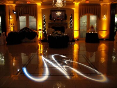 Elegant NY ballroom with warm yellow lighting, a chandelier, decorated tables, wedding cake, and white floral centerpiece. A glowing abstract light pattern is projected onto the floor—perfect for DJ services Suffolk and Nassau County.