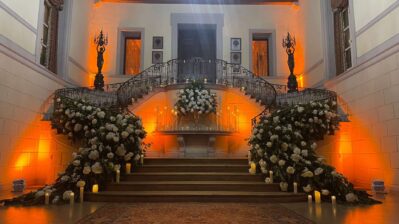A grand staircase in NY, decorated with lush white flower arrangements and candles on each side, leads to a landing adorned with more flowers. Warm orange lighting highlights the elegant, formal setting—perfect for entertainment services Suffolk and Nassau County, NY.
