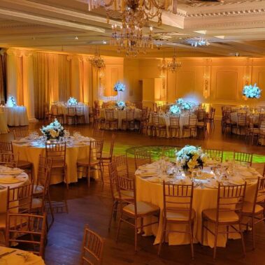 Elegant banquet hall in NY, NY decorated for an event, featuring round tables with white tablecloths, gold chairs, and grand white floral centerpieces, with warm lighting and chandeliers—ideal for entertainment services in Suffolk and Nassau County.