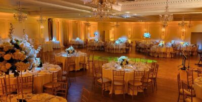 Elegant banquet hall in NY, NY decorated for an event, featuring round tables with white tablecloths, gold chairs, and grand white floral centerpieces, with warm lighting and chandeliers—ideal for entertainment services in Suffolk and Nassau County.