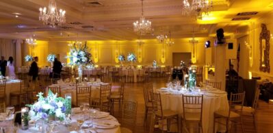 Elegant ballroom set for a formal event, featuring round tables with white linens, floral centerpieces, gold chairs, chandeliers, and warm yellow lighting. A few people are present as top DJ services Suffolk and Nassau County, NY enhance the spacious atmosphere.