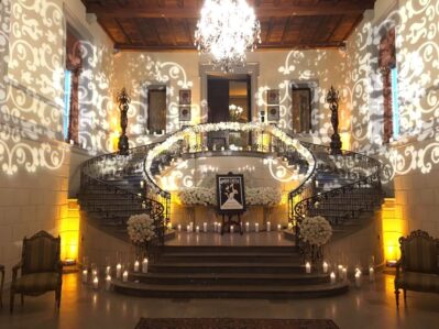 A grand staircase in NY decorated with white flowers, glowing candles, and intricate light patterns leads up to a chandelier-lit landing—perfect for events seeking top entertainment services Suffolk and Nassau County.