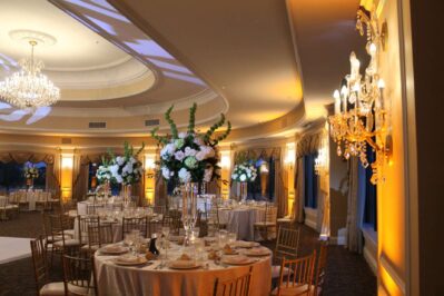 Elegant banquet hall in NY with round tables, tall floral centerpieces, gold chairs, and lit chandeliers. Warm yellow lighting adds a cozy touch—perfect for events with top entertainment services Suffolk and Nassau County.