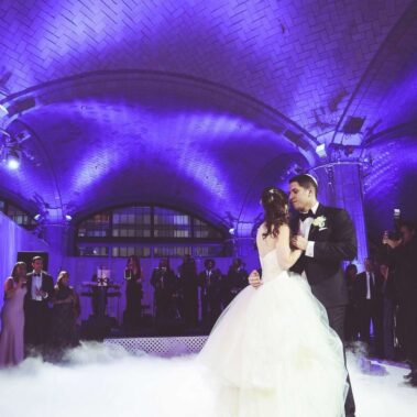 A bride and groom share their first dance under a purple-lit vaulted ceiling in NY, surrounded by guests in formal attire. A band from top entertainment services Suffolk and Nassau County, NY performs as mist covers the dance floor.