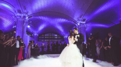 A bride and groom share their first dance under a purple-lit vaulted ceiling in NY, surrounded by guests in formal attire. A band from top entertainment services Suffolk and Nassau County, NY performs as mist covers the dance floor.
