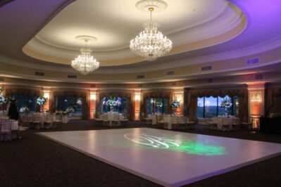 A large, elegant ballroom with chandeliers, draped windows, round dining tables, and a square white dance floor—illuminated by soft, colorful lighting and a decorative monogram projection—perfect for entertainment services in Suffolk and Nassau County.
