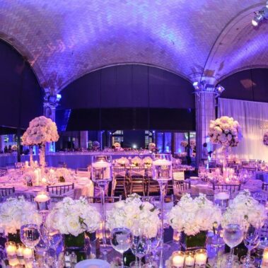 Elegant banquet hall decorated for an event with round tables, white floral centerpieces, glassware, and candles; purple and blue ambient lighting enhances the arched ceiling—perfect for entertainment services Suffolk and Nassau County, NY.