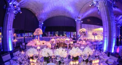 Elegant banquet hall decorated for an event with round tables, white floral centerpieces, glassware, and candles; purple and blue ambient lighting enhances the arched ceiling—perfect for entertainment services Suffolk and Nassau County, NY.