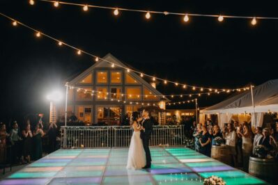 A bride and groom share a first dance on a glowing, colorful dance floor outdoors at night, surrounded by wedding guests and string lights—a magical scene made possible by top entertainment services Suffolk and Nassau County, NY. A large, lit-up house stands in the background.