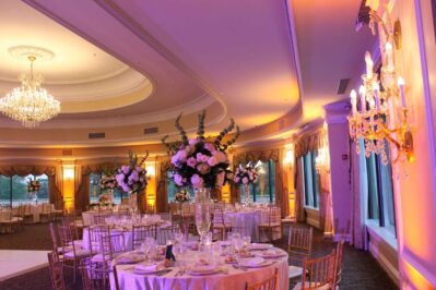 A lavishly decorated banquet hall with round tables, elegant floral centerpieces, gold chairs, chandeliers, and warm purple-yellow lighting—perfect for celebrations enhanced by top entertainment services in Suffolk and Nassau County.