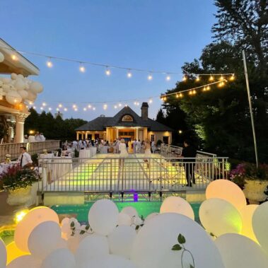 An elegant outdoor evening event with white balloons, string lights, and tables by the pool. Guests in formal attire mingle as servers attend to them, enhanced by top entertainment services Suffolk and Nassau County. Trees and a lit building are in the background.