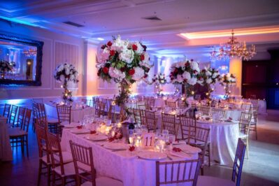 Elegant banquet hall with round tables draped in white cloths, tall floral centerpieces, and wooden chairs. Soft lighting, candles, and chandeliers set a festive mood—perfect for events with entertainment services Suffolk and Nassau County, NY.