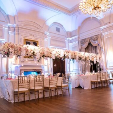 A formal banquet hall with high ceilings, chandeliers, and large paintings. Two long tables are elegantly set with tall white and pink flowers and gold chairs—perfect for events featuring DJ services in Suffolk and Nassau County, NY.