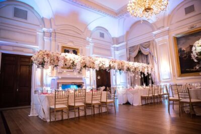 A formal banquet hall with high ceilings, chandeliers, and large paintings. Two long tables are elegantly set with tall white and pink flowers and gold chairs—perfect for events featuring DJ services in Suffolk and Nassau County, NY.