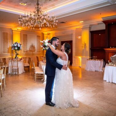 A bride and groom share their first dance in an elegant, softly lit NY ballroom with chandeliers, floral centerpieces, and white tablecloths, creating a romantic atmosphere—perfect for entertainment services Suffolk and Nassau County couples adore.