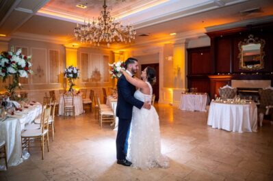 A bride and groom share their first dance in an elegant, softly lit NY ballroom with chandeliers, floral centerpieces, and white tablecloths, creating a romantic atmosphere—perfect for entertainment services Suffolk and Nassau County couples adore.