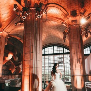A bride in a white gown stands holding a bouquet in a grand, elegant hall with tall columns and arched ceilings. Warm orange lighting highlights the details, perfect for DJ services Suffolk and Nassau County, NY to create an unforgettable celebration.