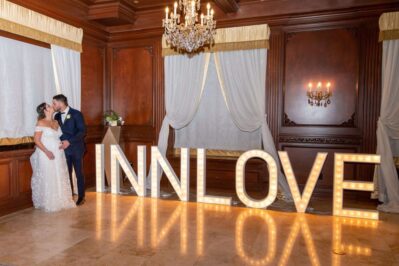 A bride and groom share a kiss in an elegant room with wood paneling and chandeliers. Large illuminated letters spell out “INN LOVE” on the polished floor, highlighting top entertainment services Suffolk and Nassau County, NY has to offer.