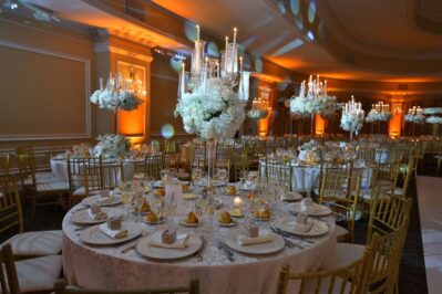 A luxurious banquet hall in NY, NY set for an event, with round tables covered in white linens, gold chairs, elegant centerpieces of white flowers, and tall candelabras; perfect for entertainment services in Suffolk and Nassau County.