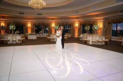 A bride and groom stand together on a white dance floor in an elegant, spacious ballroom decorated with gold chairs, round tables, tall floral centerpieces, and a chandelier overhead. Their initials are illuminated on the floor—dj services Suffolk and Nassau County, NY.