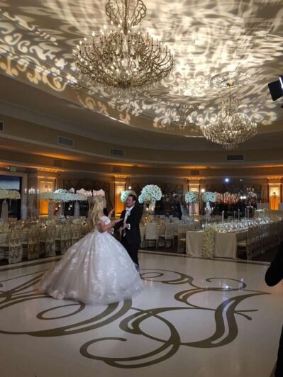 A bride and groom dance in an elegant ballroom with ornate chandeliers, decorated tables, and dramatic lighting—an unforgettable moment made even more magical by premier DJ services Suffolk and Nassau County, NY.