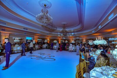 A large, elegant ballroom decorated with chandeliers and round tables filled with guests. People in formal attire are mingling and dancing on a white floor with writing on it under blue lighting, enjoying top DJ services Suffolk and Nassau County.