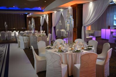 Elegant banquet hall in NY with round tables covered in white tablecloths and chairs draped in covers, set for a formal event. Tall candle centerpieces and soft lighting create the perfect setting for entertainment services Suffolk and Nassau County, NY.