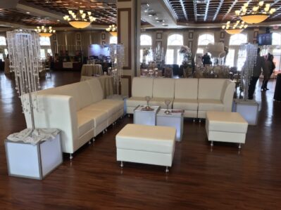 White modern sectional sofas, ottomans, and illuminated cube tables create an inviting lounge area on wooden flooring. Ideal for entertainment services in Suffolk and Nassau County, NY, the space features crystal beaded decor and elegant chandeliers above.