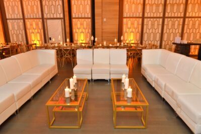 Modern lounge setup with white couches around glass coffee tables topped with candles; a long dining table and chairs with more candles are in the background—all perfect for entertainment services Suffolk and Nassau County events, in a warmly lit room.
