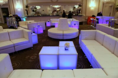 A modern lounge in NY, NY, featuring white sofas, glowing blue-lit tables, and floral centerpieces. People mingle at the bar beneath chandeliers, creating an elegant atmosphere perfect for entertainment services in Suffolk and Nassau County.