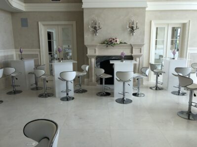A modern, elegant room in NY,NY with white high chairs and square tables set before a fireplace. Minimalist decor features flowers on the mantel and wall sconces above—perfect for entertainment services in Suffolk and Nassau County.