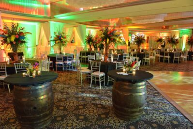 A banquet hall decorated with colorful lighting, round tables with black tablecloths, silver chairs, and large floral centerpieces. Two wooden barrel tables sit in front, creating a festive and elegant setting—perfect for DJ services in Suffolk and Nassau County, NY.