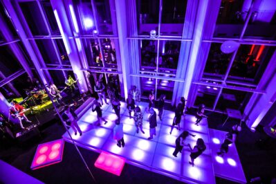 A group of people dance on a glowing, purple-lit floor in a modern venue with large windows; a live band performs under vibrant lighting—an example of top-tier entertainment services in Suffolk and Nassau County, NY.