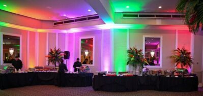 A banquet hall with colorful uplighting in green, red, and purple sets the scene. Buffet tables with black tablecloths hold food and plates as staff members stand ready. Tall floral arrangements decorate this NY,NY event by entertainment services Suffolk and Nassau County.