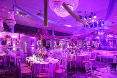 A lavish banquet hall in NY, decorated with purple and pink lighting, elegant chandeliers, draped fabric, and round tables set with floral centerpieces—perfect for entertainment services in Suffolk and Nassau County, NY.
